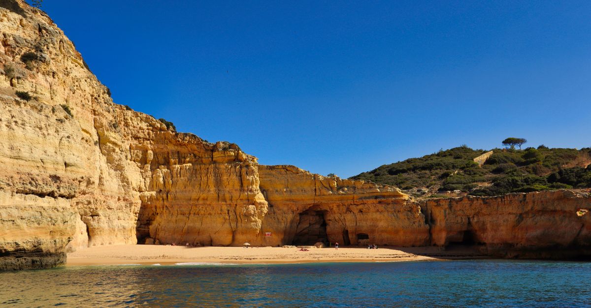 Golden limestone cliffs with a cave entrance above a narrow sandy beach near Carvoeiro, seen from the water
