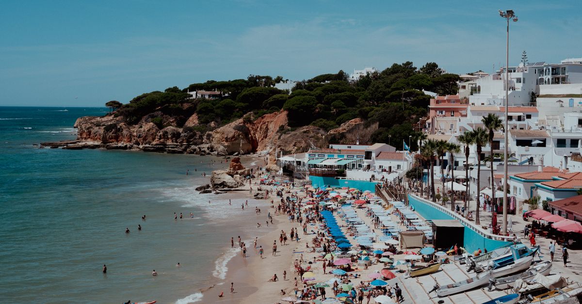 Busy sandy beach with colourful parasols and rows of sunbeds, fishing boats on a ramp, reddish cliffs with pine trees and white houses behind