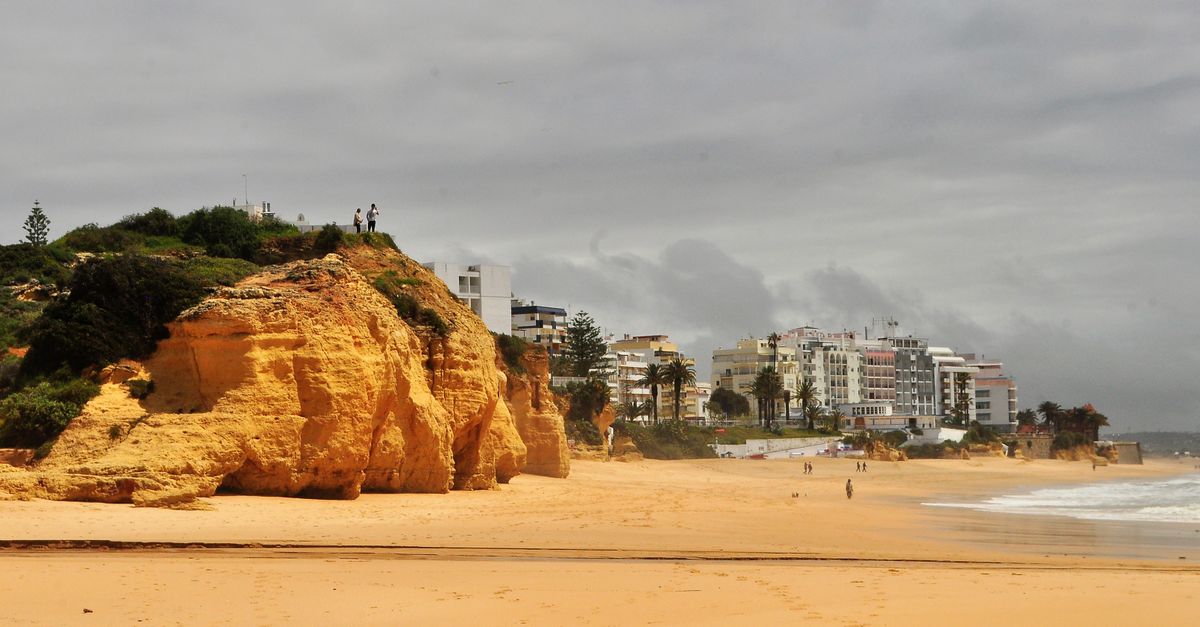 Empty sandy beach on the Algarve under a dark cloud cover, orange rock and white hotel buildings in the background